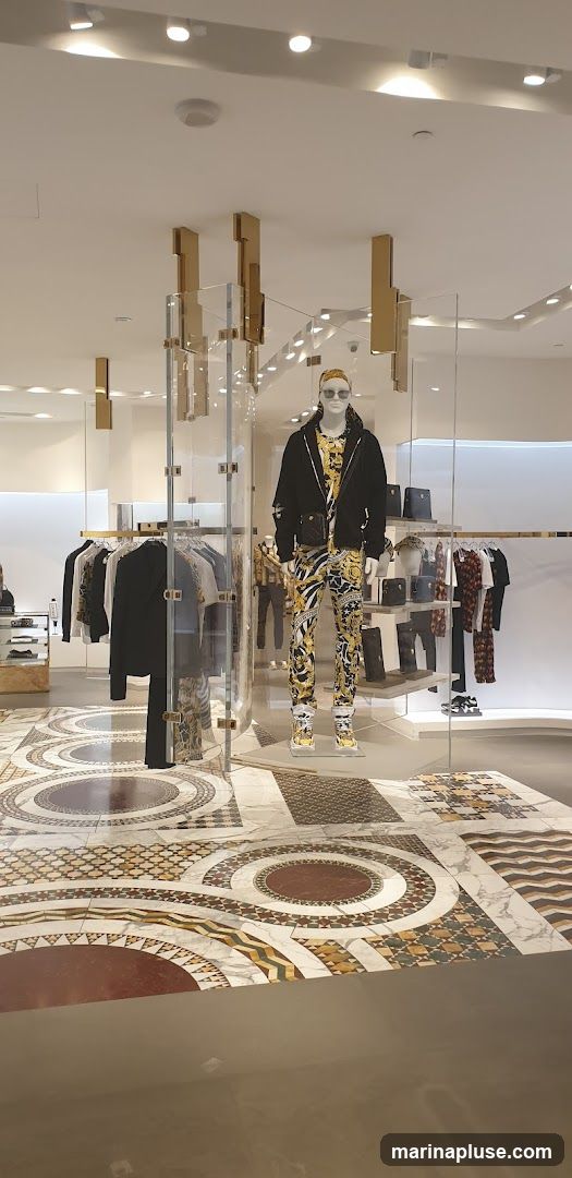A well-lit interior of a high-end fashion store. A mannequin is prominently displayed wearing a black jacket over a patterned shirt and matching patterned pants, accessorized with sunglasses and a small crossbody bag. To the left, racks of clothing are visible, including black jackets and patterned shirts. Shelves in the background hold handbags and other accessories. The floor features an intricate marble mosaic design.