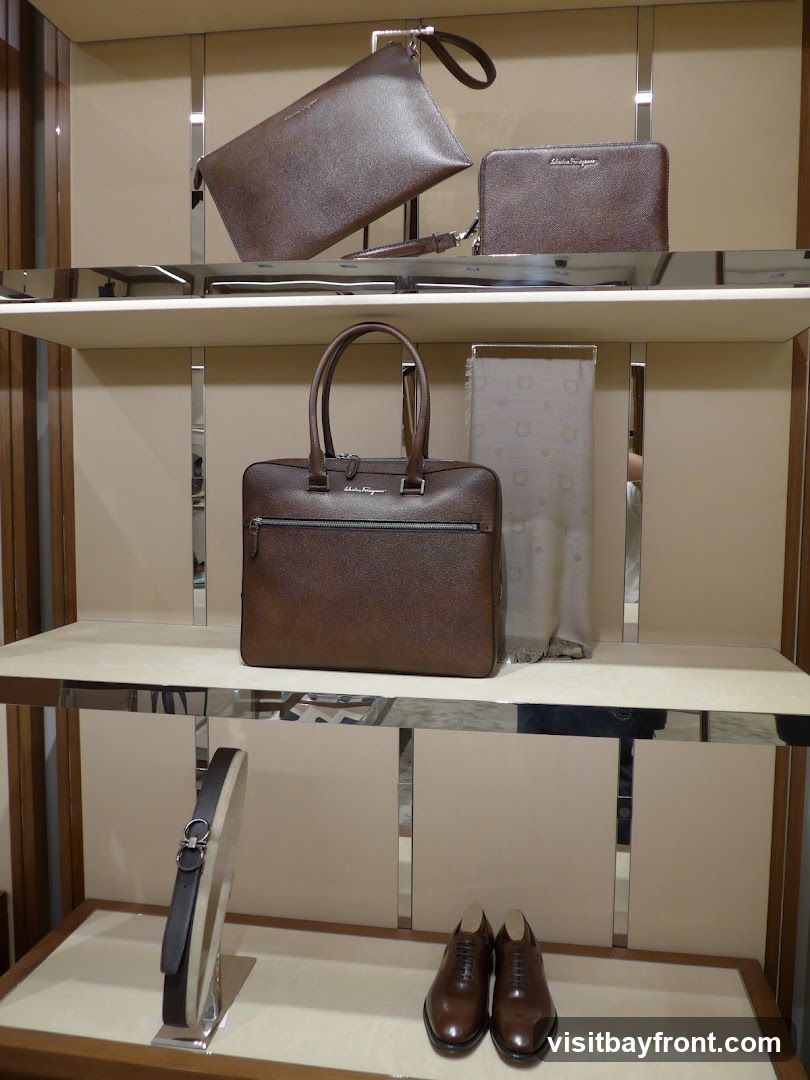 A display of luxury leather goods, including handbags, a clutch, a wallet, a scarf, a belt, and dress shoes, arranged on shelves in a store. The items are predominantly brown and made of textured leather. The brand name "Salvatore Ferragamo" is visible on the handbag and wallet.