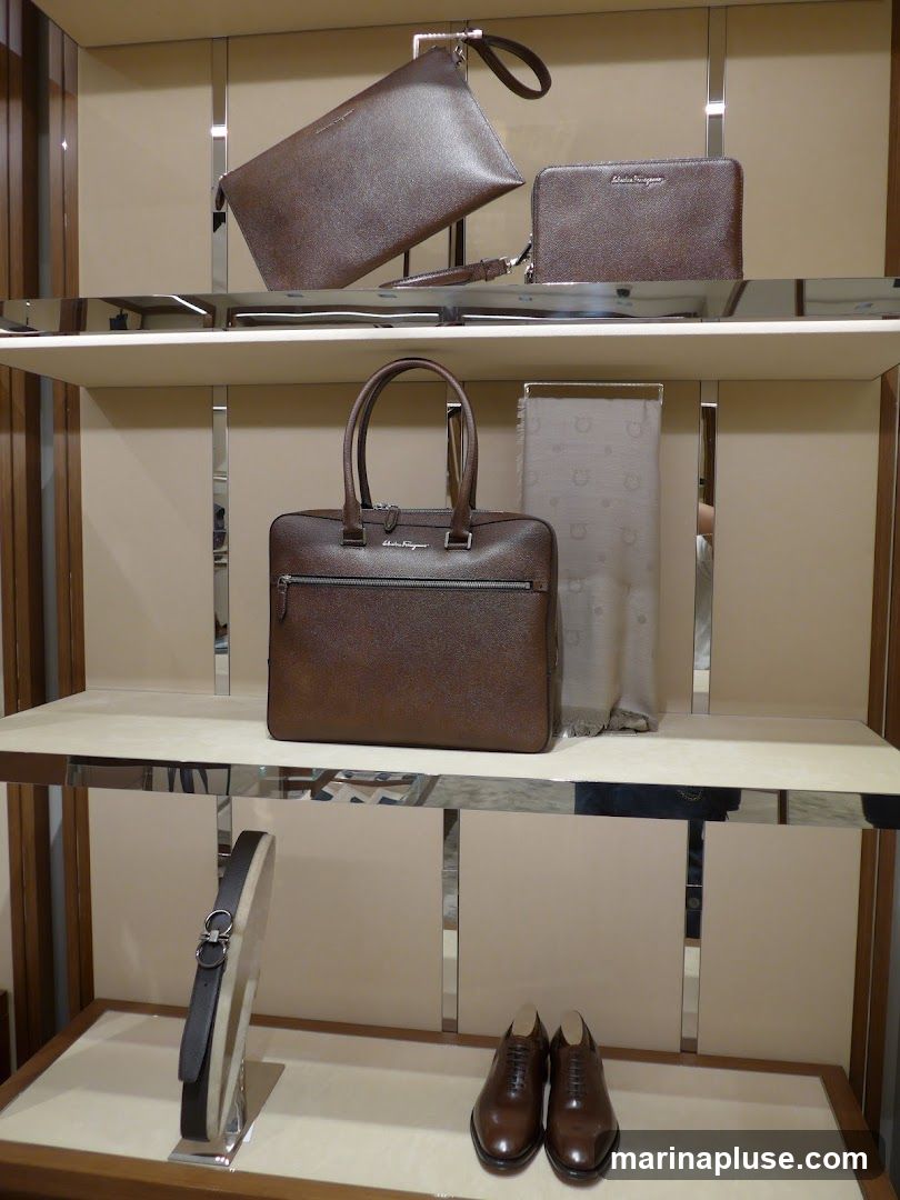 A display of luxury leather goods, including handbags, a clutch, a wallet, a scarf, a belt, and dress shoes, arranged on shelves in a store. The items are predominantly brown and made of textured leather. The brand name "Salvatore Ferragamo" is visible on the handbag and wallet.