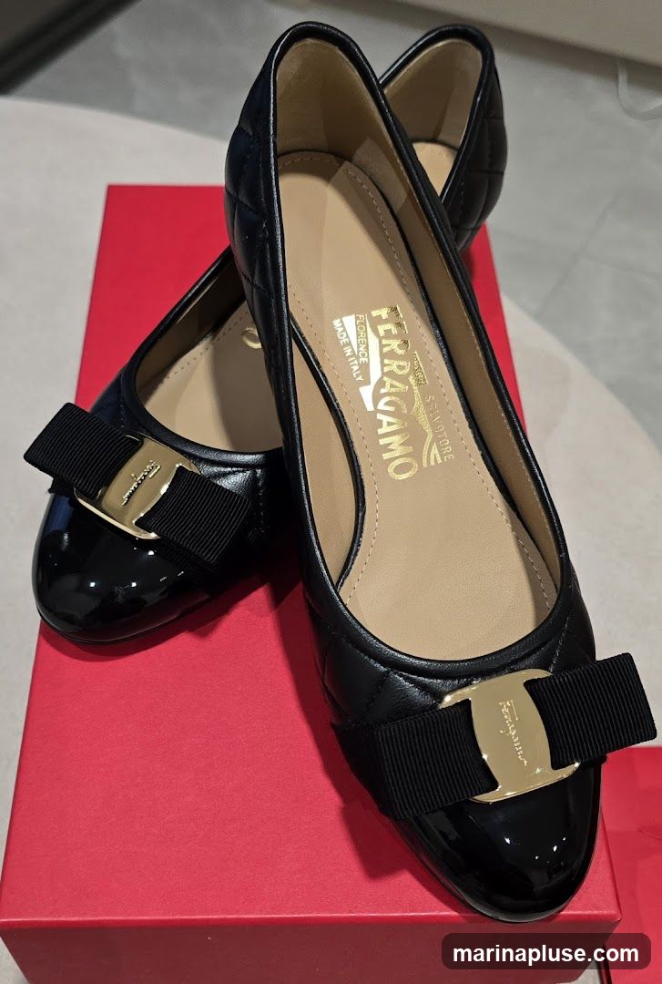 A pair of black quilted leather ballet flats with a patent leather toe cap and a black grosgrain ribbon bow with a gold Gancini buckle. The shoes are resting on a red box. The brand name "FERRAGAMO" and "SALVATORE FERRAGAMO" are visible on the insoles, along with "FLORENCE" and "MADE IN ITALY".