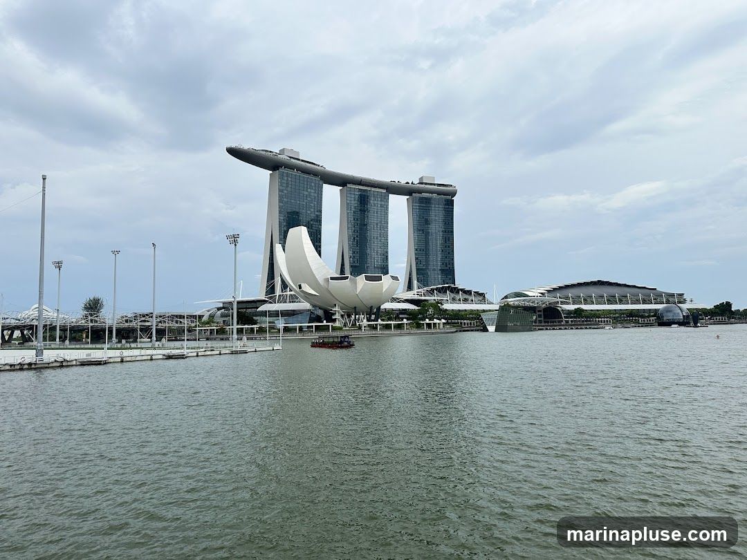 A wide shot of the Marina Bay Sands hotel and the ArtScience Museum in Singapore, viewed across a body of water. The iconic three towers of the hotel are visible, topped with the ship-like structure. In front of the hotel, the lotus-shaped ArtScience Museum is prominent. The water in the foreground is choppy under a cloudy sky. A small boat is navigating the water between the museum and the hotel. In the background, other modern buildings and structures are visible along the waterfront.
