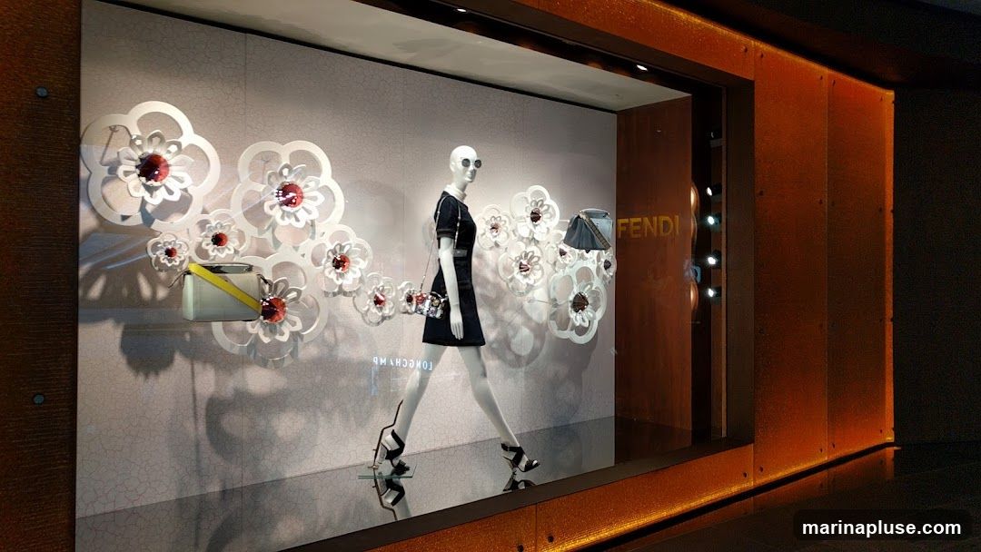 A Fendi store window display features a mannequin dressed in a black outfit, wearing sunglasses and high heels. The mannequin is adorned with a patterned handbag and is positioned among large white floral cutouts with red centers. Two handbags are also displayed, one on the left with a yellow strap and another on the right. The Fendi logo is visible in gold lettering on the right side of the display. The storefront has a warm, orange-toned exterior.