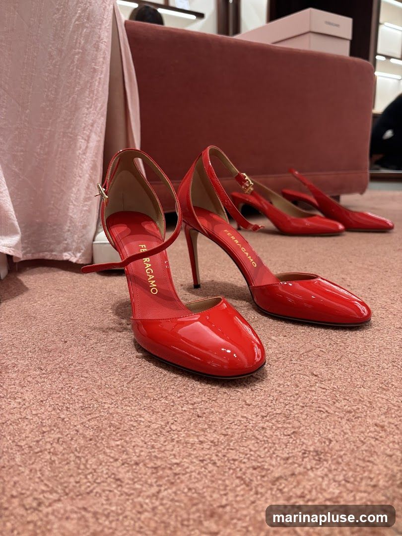 A close-up shot of several pairs of shiny red patent leather high-heeled shoes displayed on a pink carpet. The shoes have ankle straps and pointed toes. The brand name "FERRAGAMO" is visible on the insole of one of the shoes. In the background, there are pink upholstered benches and boxes, suggesting a retail environment.