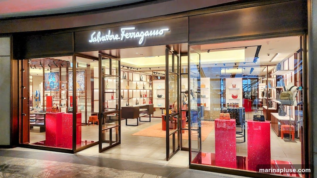 The exterior of a Salvatore Ferragamo store. The entrance features large glass doors and windows displaying various handbags and accessories. The store's name is illuminated in white script above the entrance. Inside, shelves are stocked with merchandise, and red display stands are prominent.