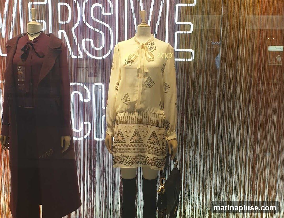 A fashion display window features two mannequins dressed in elegant clothing. The mannequin on the left wears a deep red coat with a bow tie, while the mannequin on the right is adorned in a cream-colored dress with intricate patterns and a bow at the neck. Both mannequins are accessorized with dark knee-high boots. Behind the mannequins, a neon sign with white lettering is visible, spelling out "VERSIVE CHIC". The background is a curtain of thin, vertical strands in varying shades of brown and gold, creating a textured and shimmering effect. A dark handbag is placed in front of the right mannequin.