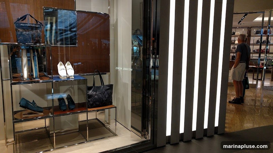 A display window of a Michael Kors store showcases various accessories. On the left, a black patterned tote bag is displayed on a shelf, along with white and blue belts. Below, a pair of blue loafers and another pair of dark blue shoes are visible. On the right side of the window, a man in a grey t-shirt and white shorts stands looking into the store. The store interior features shelves stocked with handbags. The store name "MICHAEL KORS" is visible in reverse on the glass.