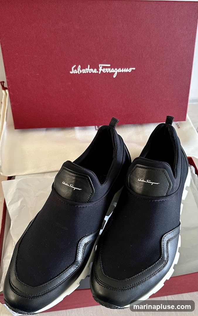 A pair of black Salvatore Ferragamo slip-on sneakers are displayed in their red branded box. The sneakers have a black fabric upper with leather accents and white soles. The Salvatore Ferragamo logo is visible on a leather patch on each shoe and on the box lid.