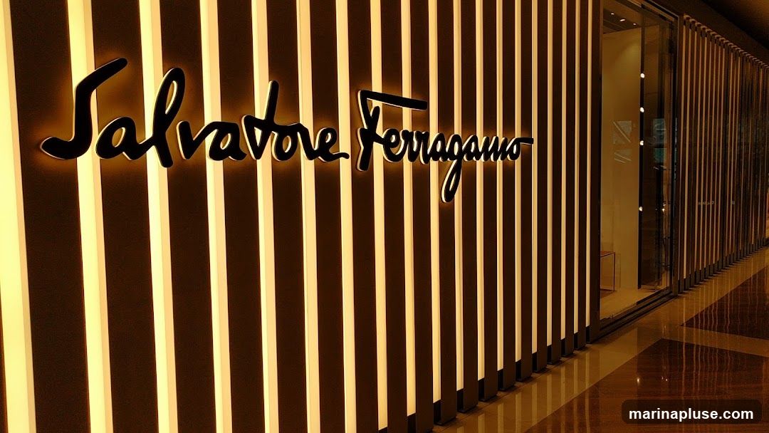 The exterior of a Salvatore Ferragamo store. The store features a prominent illuminated sign with the brand's name in a script font. The facade is decorated with vertical, backlit panels, creating a warm and inviting ambiance. A glass entrance is visible to the right, reflecting the interior lights. The floor is polished and reflects the lighting from the store and the surrounding area.
