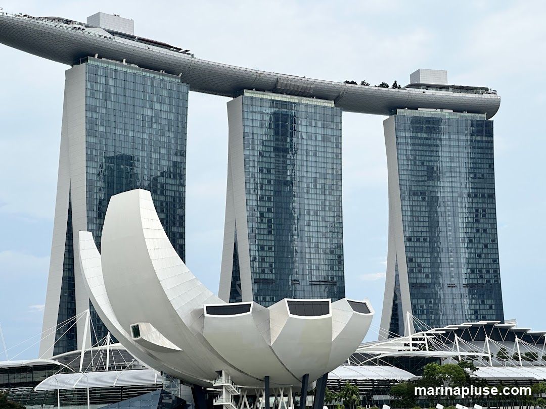 A wide shot of the Marina Bay Sands complex in Singapore, featuring its iconic three towers connected by a sky-high platform and the lotus-shaped ArtScience Museum in the foreground. The buildings are modern, with glass facades reflecting the cloudy sky.