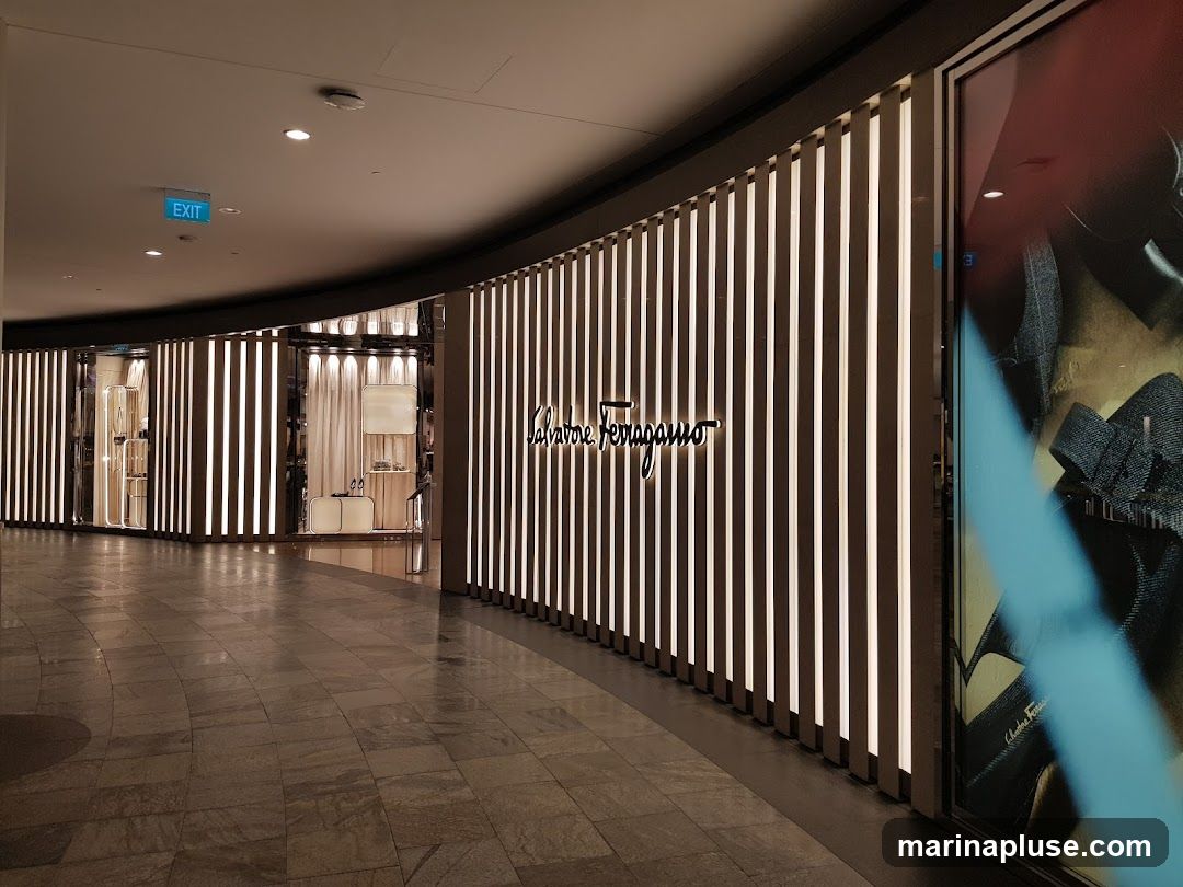 The exterior of a Salvatore Ferragamo store with vertical lighting panels and the brand's logo prominently displayed. The store features a display window on the right side with a fashion advertisement. An exit sign is visible on the ceiling.