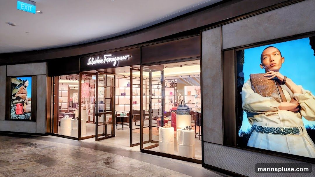 The exterior of a Salvatore Ferragamo store. The storefront features a large glass entrance with a neon sign above that reads "Salvatore Ferragamo". Inside, shelves are stocked with handbags and shoes. To the left and right of the entrance are large digital screens displaying advertisements. An "EXIT" sign is visible above the entrance.