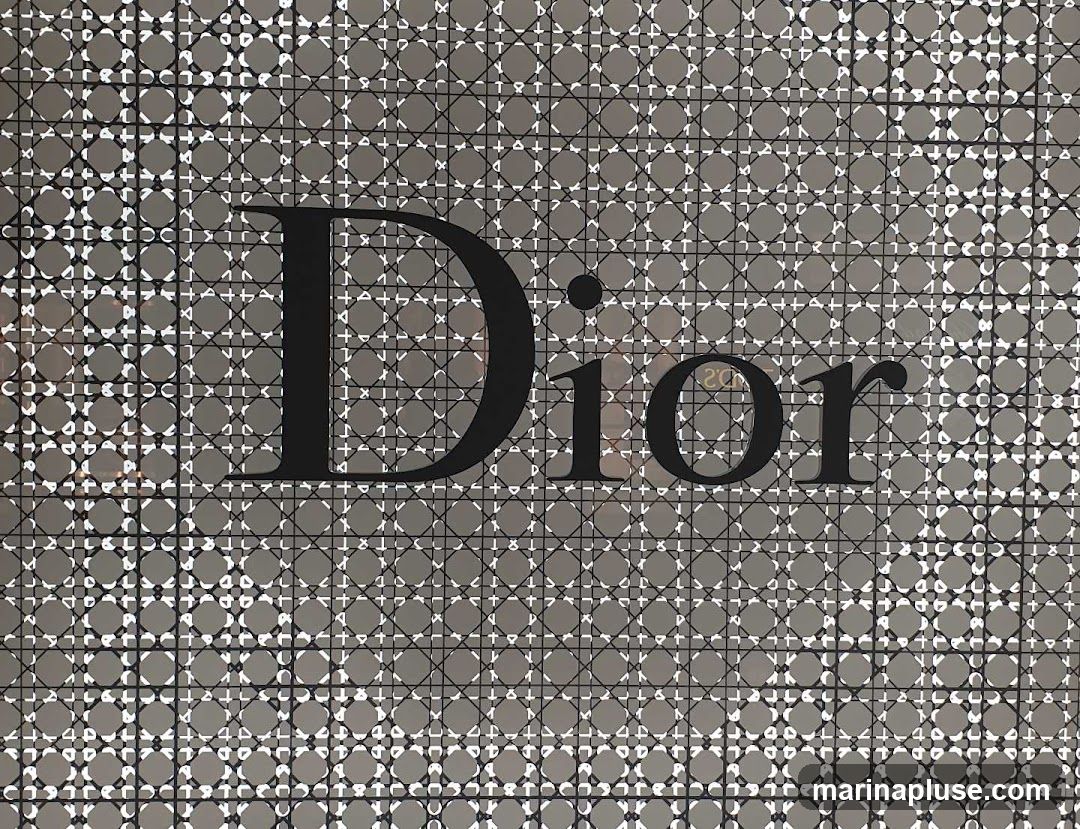 A close-up view of the Dior logo against a patterned background. The logo is in black, bold, sans-serif font. The background features a repeating geometric pattern of interlocking circles and crosses, with a subtle sheen.