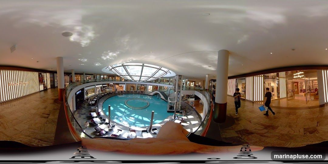 A panoramic view of a modern indoor shopping mall. In the center is a large, circular pool of blue water with a fountain. Surrounding the pool are multiple levels of shops and walkways. The shops have illuminated facades, and some display merchandise. People are seen walking through the mall and sitting at tables near the pool. The ceiling features a large, geodesic dome skylight allowing natural light to enter.