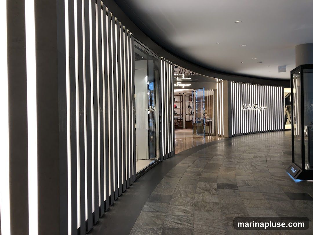 A view of the exterior of a Salvatore Ferragamo store in a modern shopping mall. The storefront features a curved facade with vertical illuminated panels and the store's logo prominently displayed. Through the glass entrance, shelves with merchandise are visible.