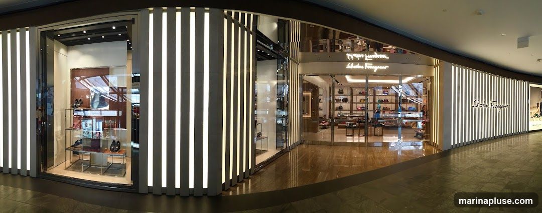 A panoramic view of the exterior of a Salvatore Ferragamo store. The facade features vertical white lighting strips and large glass windows displaying merchandise. The store's name, "Salvatore Ferragamo," is prominently displayed in script on the upper part of the building. Inside, shelves are stocked with handbags and other luxury goods.