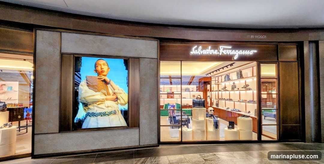 The exterior of a Salvatore Ferragamo store. A large digital screen displays a model. The store entrance features a prominent neon sign with the store's name. Inside, shelves display shoes and handbags.