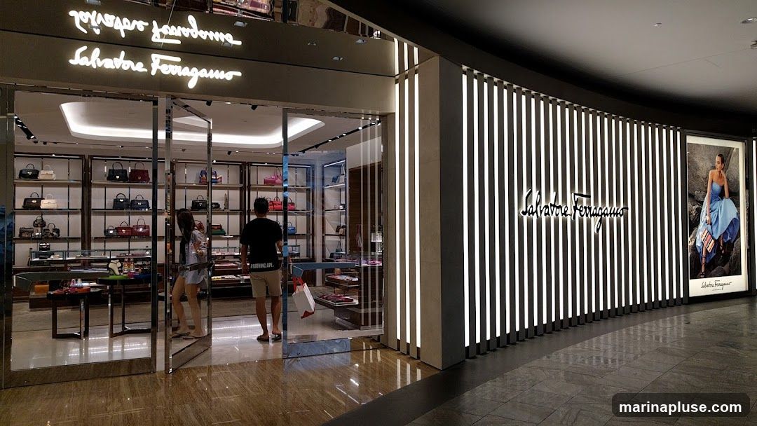 The exterior of a Salvatore Ferragamo store. The entrance is a glass door with a reflective frame. Inside, shelves display various handbags. The storefront features a large illuminated sign with the Salvatore Ferragamo logo and a vertical striped facade. A promotional poster of a woman in a blue dress is displayed on the right side.