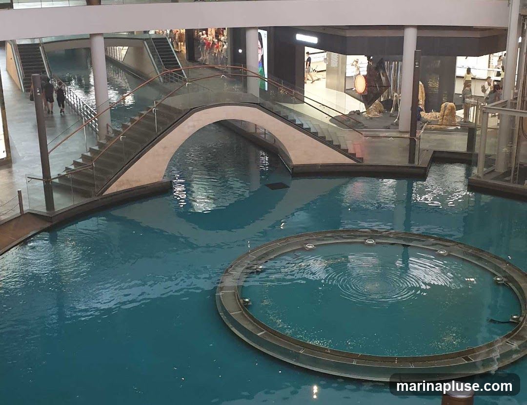 An indoor scene featuring a canal with blue water, a curved bridge with stairs, and surrounding walkways and shops. A circular fountain is visible in the foreground. People are seen walking on the upper levels and near shop entrances.