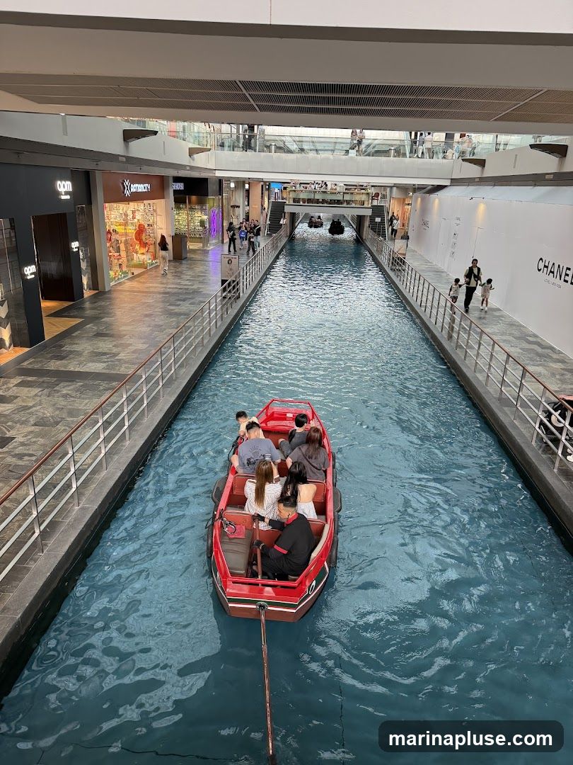 A red boat with several passengers navigates a blue indoor canal within a shopping mall. Shops are visible on either side of the canal, including one with a large "CHANEL" sign. People are walking along the walkways beside the canal.