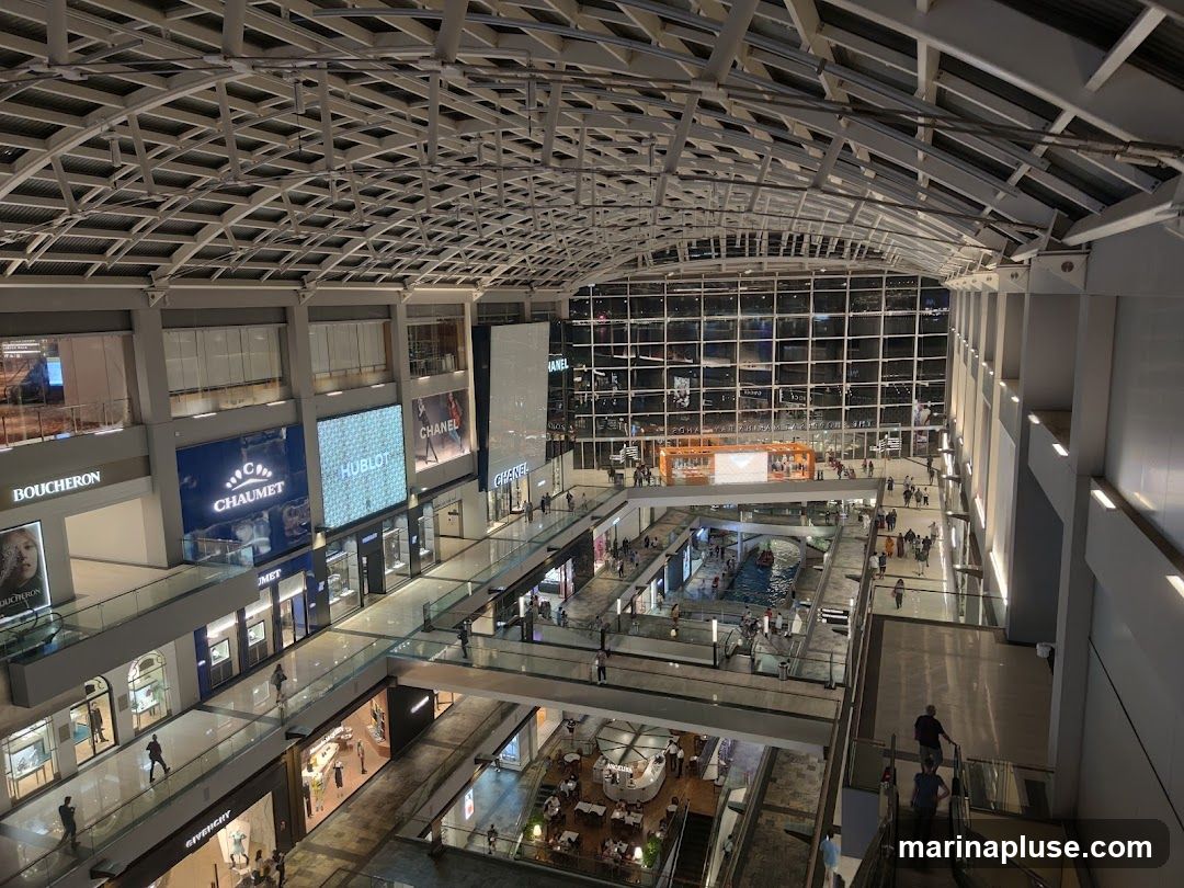 An overhead view of the interior of a modern shopping mall. The mall features multiple levels with shops, escalators, and walkways. Large digital displays show brand names like Boucheron, Chaumet, and Hublot. A large glass facade at the far end of the mall offers a view of the outside. People are seen browsing shops and walking around.