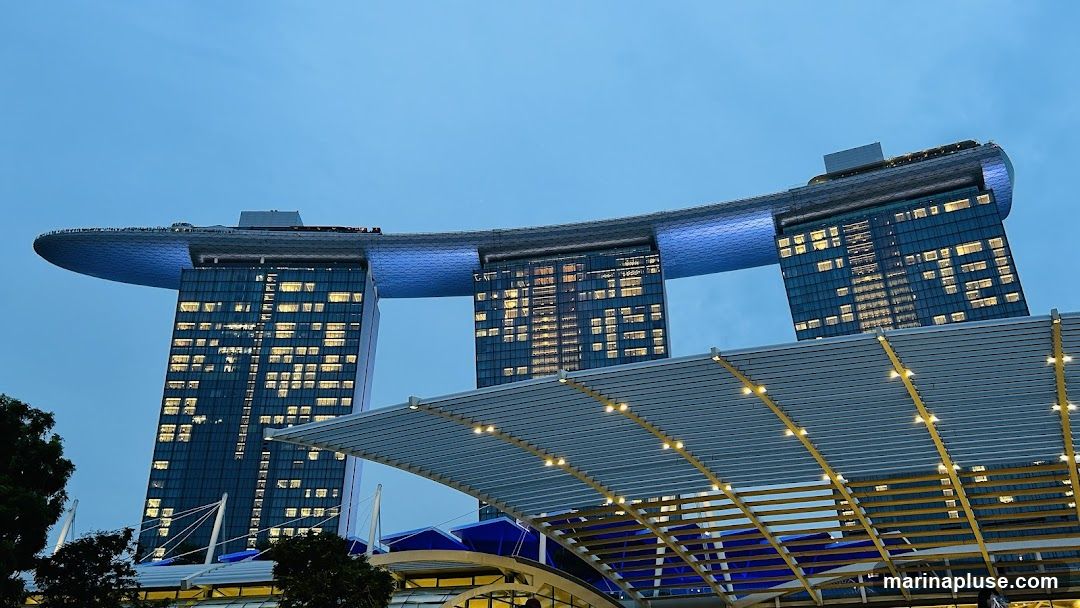 A wide shot of the Marina Bay Sands hotel in Singapore, with its iconic three towers connected by a sky park. The building is illuminated with lights, and the sky is a deep blue, suggesting dusk or early evening. The foreground shows a modern architectural structure with a canopy and lights.