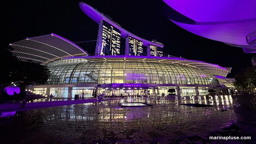 A wide shot of a large, modern building complex at night, illuminated with purple lights. The building features a distinctive curved glass facade and a multi-tiered roof structure. In the foreground, a reflective pool mirrors the building's lights and architecture. Several people are visible walking around the entrance area. The iconic Marina Bay Sands hotel towers are visible in the background, also lit with purple lights.