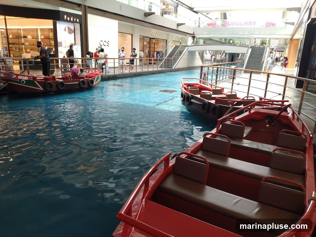 Several red passenger boats are docked on a bright blue indoor canal, likely within a shopping mall. In the background, shops with visible signage like 'Pedro' and 'COS' line the canal. A 'BURBERRY' banner is visible on an upper level. People are seen walking along the canal's edge and boarding one of the boats. The scene suggests a recreational boat ride attraction within a commercial space.