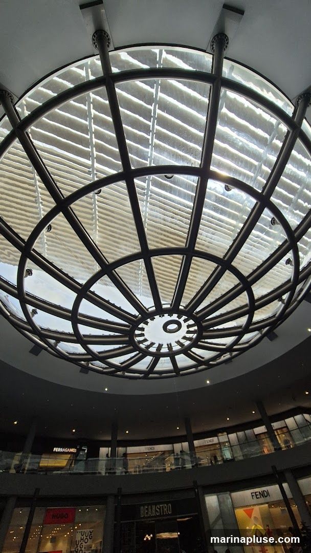 The image shows the interior of a shopping mall with a large, circular, domed ceiling made of glass and metal. Below the dome, multiple levels of shops are visible, with prominent signage for brands like FERRAGAMO, FENDI, and HUGO. A restaurant or cafe named BEANSTRO is also visible. The overall impression is of a modern, upscale retail environment.