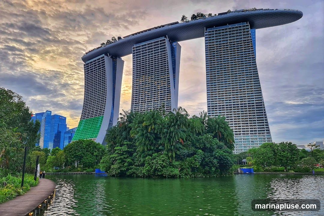 A wide shot of the Marina Bay Sands hotel in Singapore, with its distinctive three towers connected by a sky-high platform. The hotel is set against a dramatic sky with clouds catching the golden light of either sunrise or sunset. In the foreground, a calm green body of water reflects the sky and the surrounding lush greenery. A wooden boardwalk curves along the edge of the water, leading towards the hotel. Other modern skyscrapers are visible in the background.
