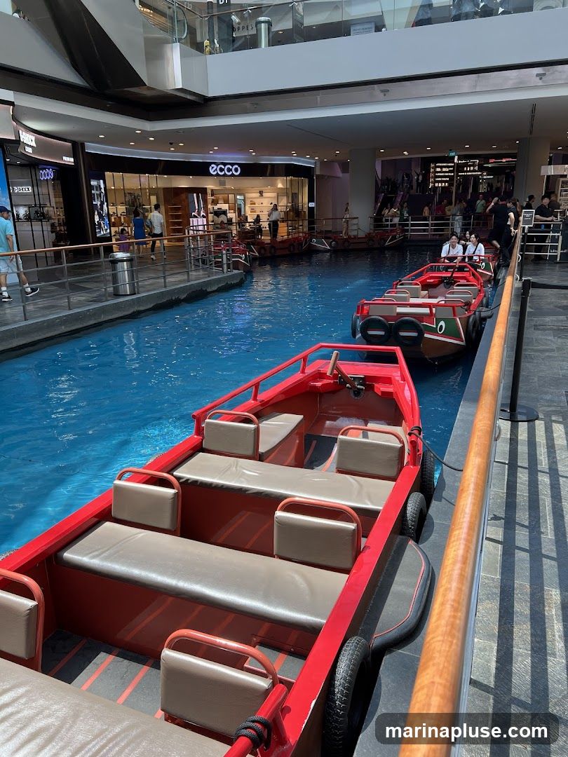 Red boats with tan seats are lined up in a bright blue artificial canal within a modern indoor shopping mall. Shops with illuminated signage, including 'ECCO', are visible in the background. People are seen walking along the canal's edge and riding in some of the boats.
