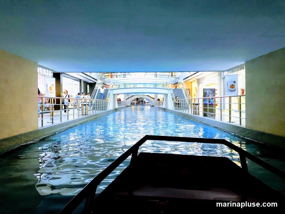 A view from a boat on an indoor canal, with shops and escalators visible on either side. The water reflects the bright lights of the shopping mall.