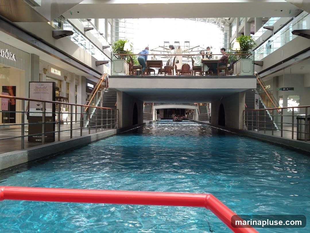An indoor canal with bright blue water flows through a modern shopping mall. Gondolas are visible in the distance. Shops line the upper levels of the mall, with people dining at tables on a balcony overlooking the canal. A red railing is in the foreground.