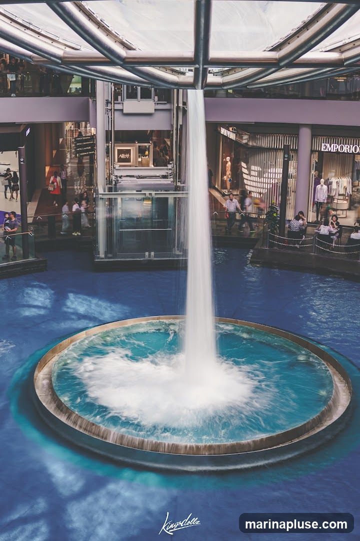 An indoor shopping mall features a large, circular fountain with a powerful waterfall cascading into it. Surrounding the fountain are multiple levels of shops, with visible storefronts and mannequins displaying clothing. People are seen walking on the elevated walkways and sitting at tables near the shops. The architecture includes a modern, metallic ceiling structure.