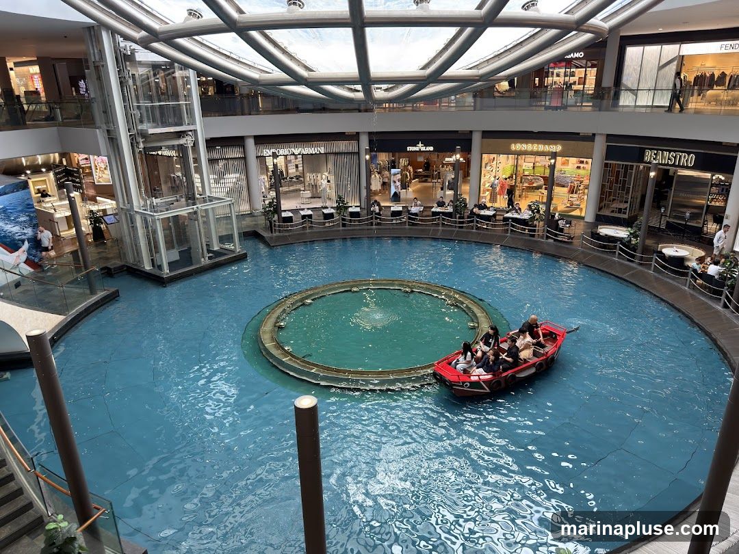 An indoor canal with a red boat carrying passengers, surrounded by shops and restaurants on multiple levels. The canal has a circular fountain in the center. Shops visible include Emporio Armani, Fendi, Stone Island, Longchamp, and Beanstro.