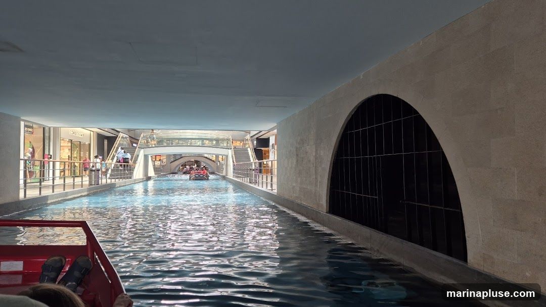 A view from a red boat on an indoor canal, with shops lining one side and arched architectural features on the other. Several gondola-like boats are visible in the distance, carrying passengers. People are seen walking along the elevated walkway next to the shops. The scene appears to be within a shopping mall or entertainment complex designed to resemble Venice.