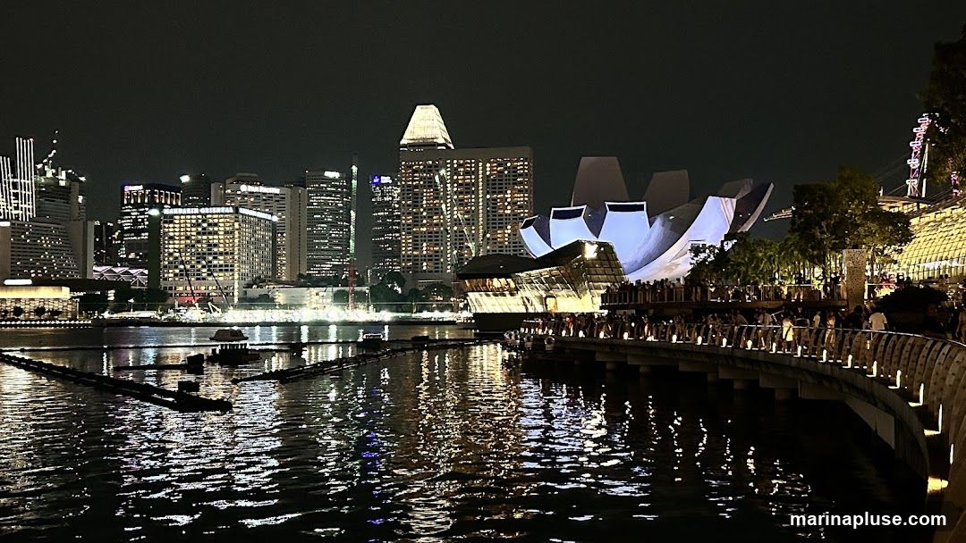 A nighttime view of a modern cityscape with illuminated skyscrapers and a distinctive lotus-shaped building. A waterfront promenade is filled with people, and the lights of the city are reflected in the dark water.