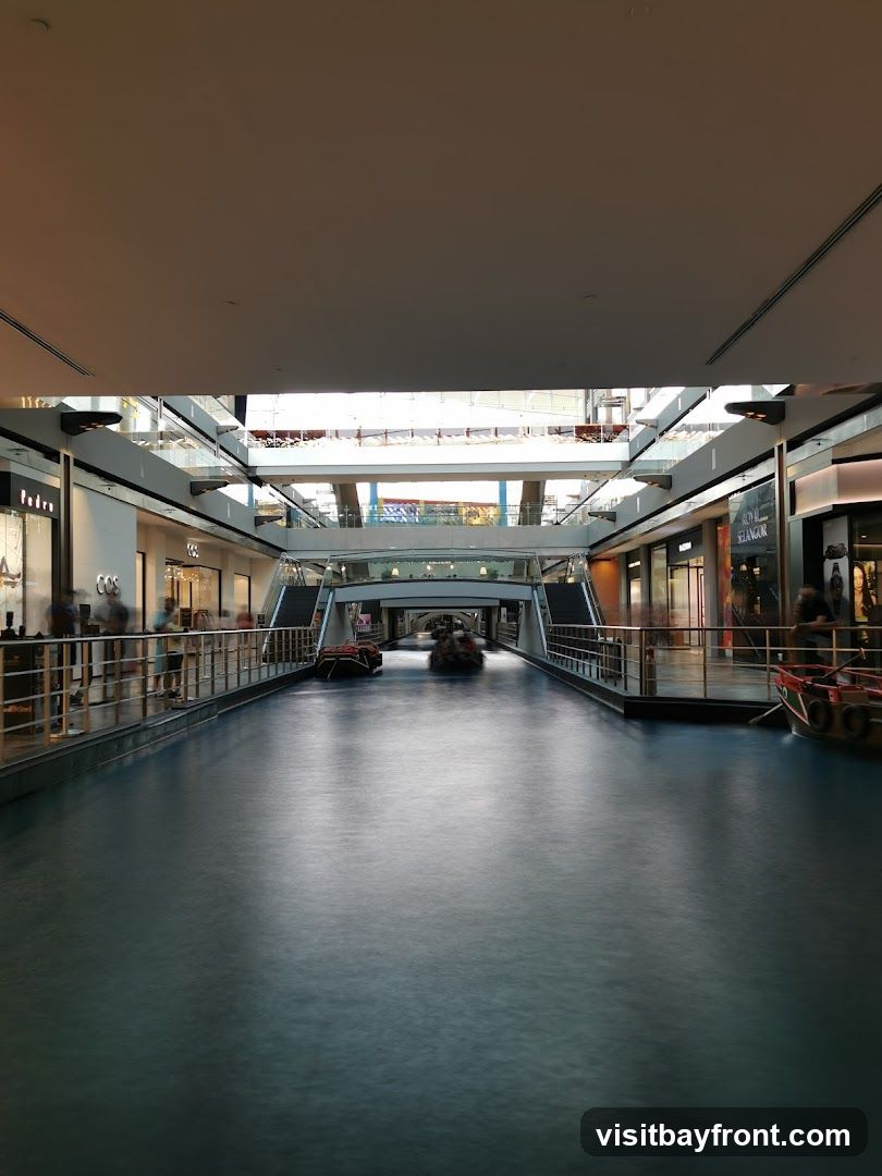 An indoor canal with boats sailing on it, surrounded by shops on multiple levels. The architecture suggests a modern shopping mall with a unique water feature. People are visible walking along the elevated walkways and near the shops.