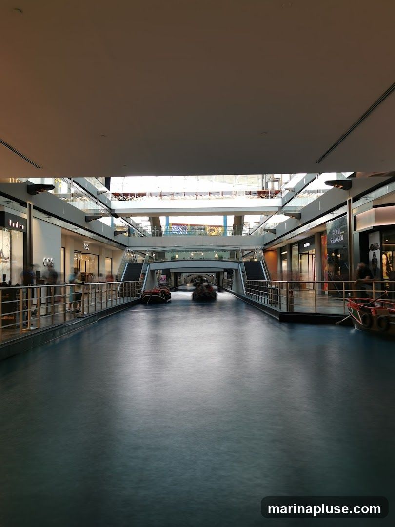 An indoor canal with boats sailing on it, surrounded by shops on multiple levels. The architecture suggests a modern shopping mall with a unique water feature. People are visible walking along the elevated walkways and near the shops.