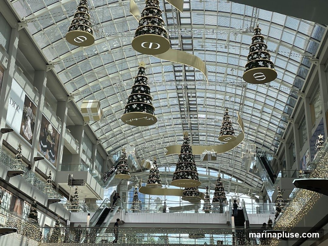A view of a mall interior decorated for Christmas with tiered Christmas trees hanging from the ceiling. The trees are adorned with stars and lights, and some have large circular ornaments with the Dior logo. Large advertisements for Dior are visible on the walls. Escalators and shoppers can be seen on multiple levels of the mall. The ceiling is made of glass panels, allowing natural light to filter in.
