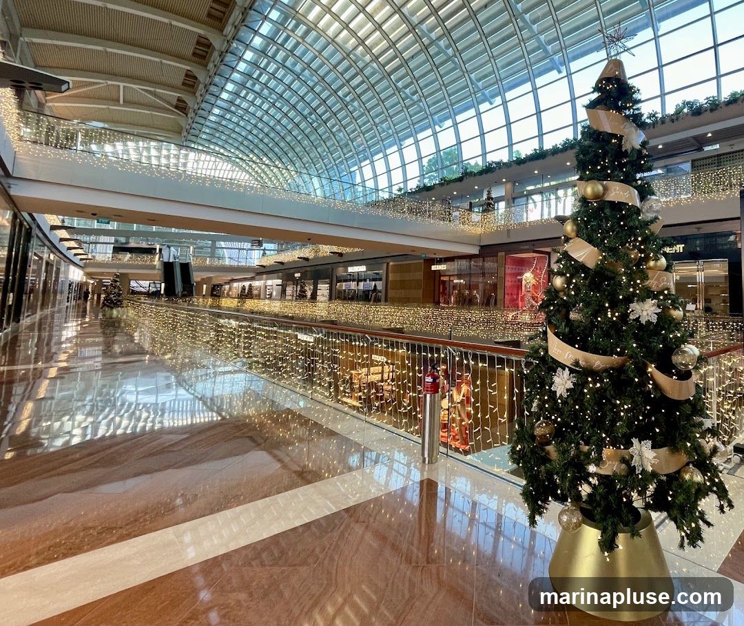 A wide-angle view of a modern shopping mall interior, adorned with festive Christmas decorations. A large, decorated Christmas tree stands prominently in the foreground on the right. The mall features multiple levels, with escalators visible in the distance. The walkways and railings are extensively decorated with strings of warm white fairy lights, creating a magical ambiance. The ceiling is a distinctive glass dome, allowing natural light to filter in. Shops line the corridors on both sides, with some storefronts visible in the background. The floor is polished marble with a striped pattern.