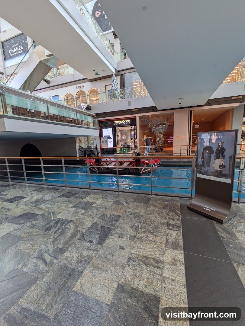 An indoor view of a shopping mall with a canal and gondola in the center. Shops like Chanel, Fred, Samsonite, and Pedro are visible on different levels. A digital display stands in the foreground with an advertisement.