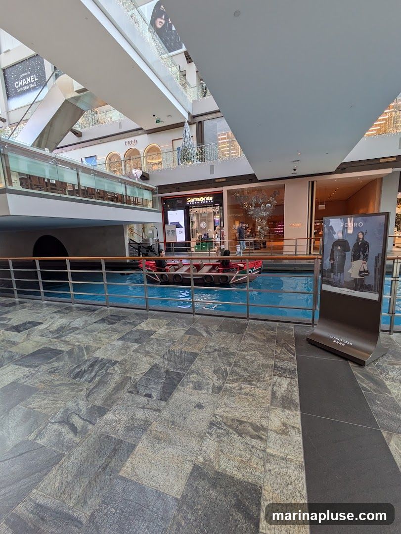 An indoor view of a shopping mall with a canal and gondola in the center. Shops like Chanel, Fred, Samsonite, and Pedro are visible on different levels. A digital display stands in the foreground with an advertisement.