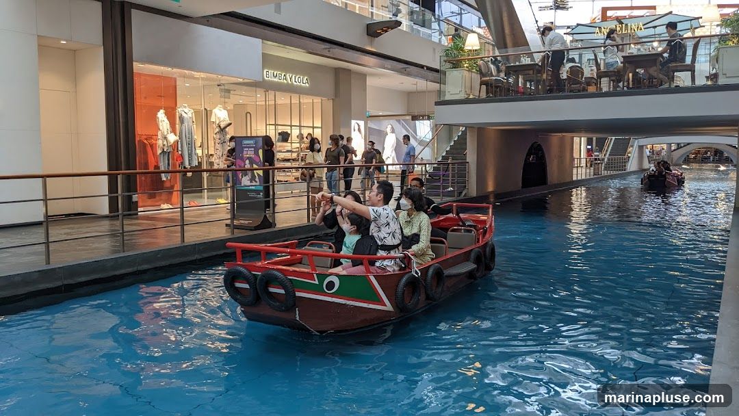 A boat carrying several people navigates a canal within a large indoor shopping mall. The boat has a red exterior with black tires along its sides. In the background, there are shops with mannequins displaying clothing, and a cafe with patrons seated at tables. Another boat is visible further down the canal.