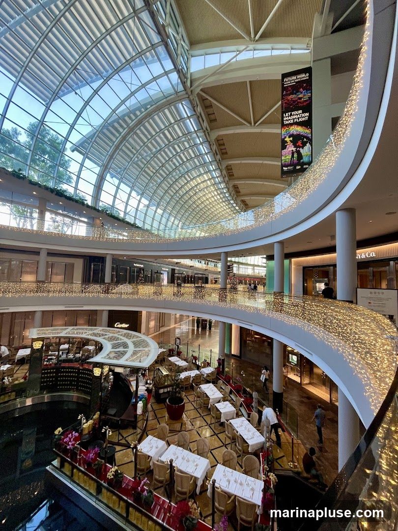 An interior view of a multi-level shopping mall with a large glass ceiling. The atrium features a central dining area with tables set for meals, surrounded by shops. Decorative lights adorn the railings of the upper levels. A banner for 'Future World' is visible hanging from the ceiling.