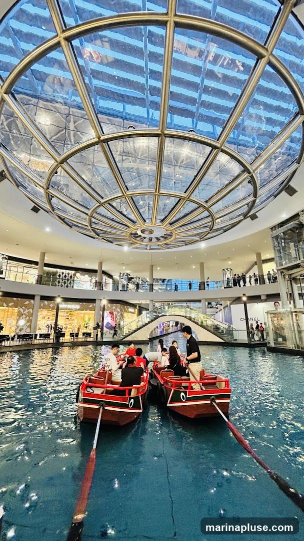 People are enjoying a gondola ride in a canal that runs through a large, multi-level shopping mall. The canal is surrounded by shops and escalators. A large, circular glass ceiling allows natural light to filter into the space.