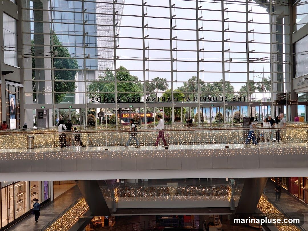 The image shows the interior of a large, modern shopping mall. A multi-level walkway is visible, adorned with strings of warm fairy lights. People are seen walking along the upper walkway, which overlooks lower levels of shops. The background features a large glass facade with a grid pattern, revealing palm trees and a building outside. The text 'THE SHOPPERS AT MARINA BAY SANDS' is visible, reflected on the glass.