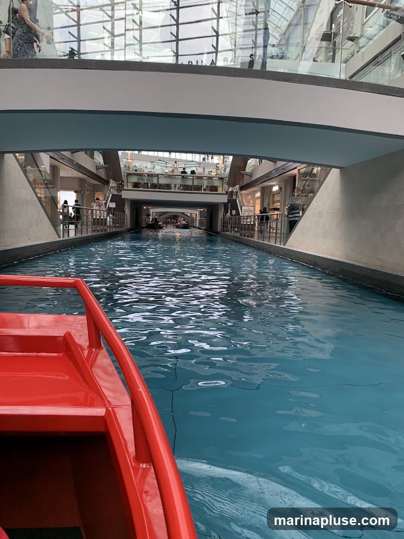 A view from a red boat on a canal inside a modern building. The canal is lined with shops and walkways, with people visible on the upper levels. The building has a glass ceiling and a contemporary architectural style.