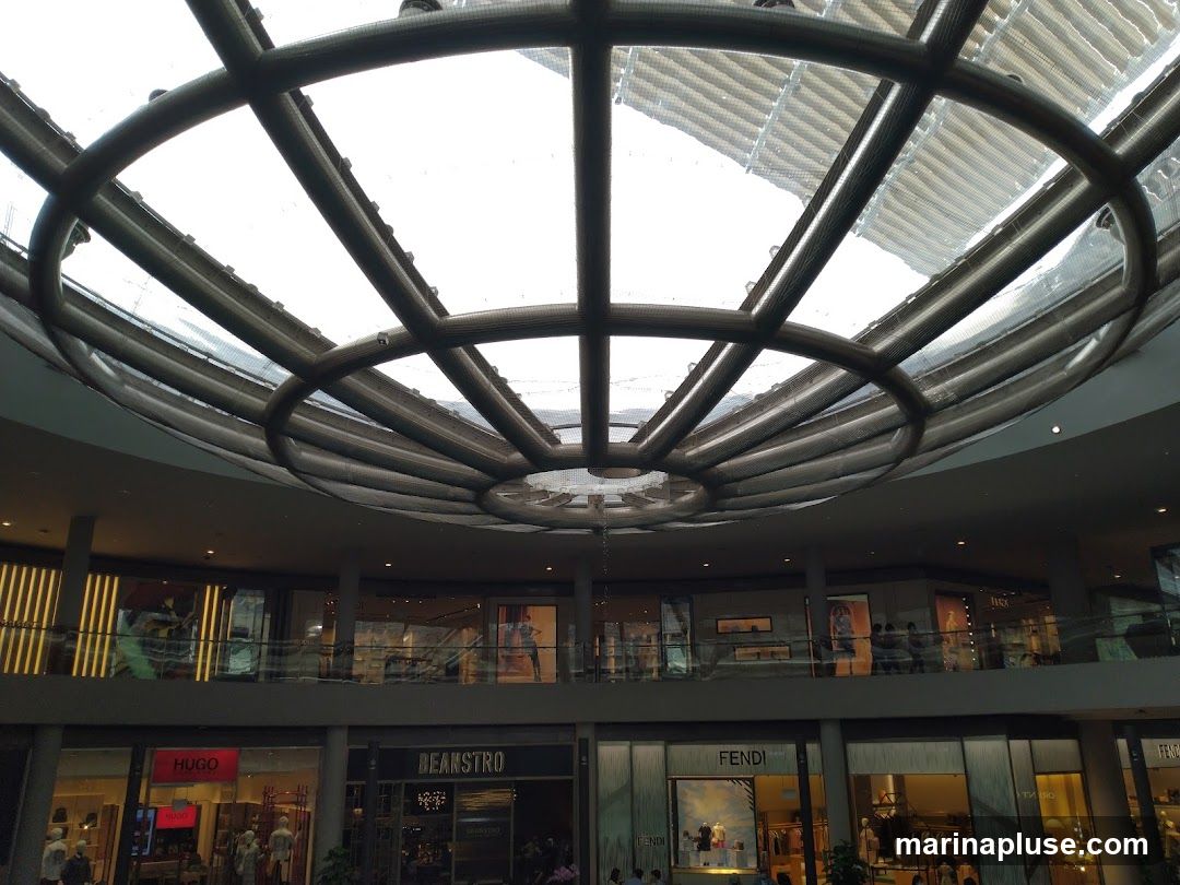 The image shows the interior of a modern shopping mall with multiple levels. A large, circular skylight with a metal framework dominates the upper portion of the image, allowing natural light to filter in. Below the skylight, various storefronts are visible on two levels. Brands such as HUGO, BEANSTRO, and FENDI are identifiable on the lower level. The upper level also features storefronts with displays and mannequins. The overall impression is one of a spacious and well-lit retail environment.