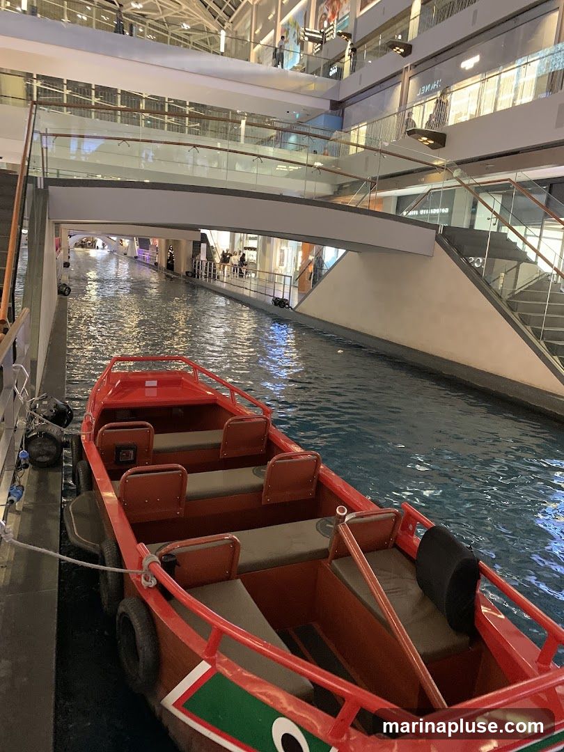 A red boat with multiple rows of seats is docked in an indoor canal. The canal is surrounded by a multi-level shopping mall with escalators and shops visible on the upper floors.