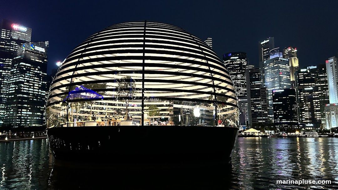 A spherical glass building with horizontal black stripes, illuminated from within, sits on the water at night. The building's interior is visible, showing trees, displays, and people. In the background, a city skyline with many illuminated skyscrapers rises against the dark sky. The water in the foreground reflects the lights of the city and the building.